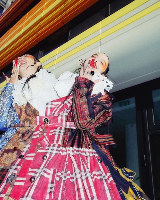 Two women in colorful, patterned dresses stand close together with smiles and exaggerated makeup against an abstract background.