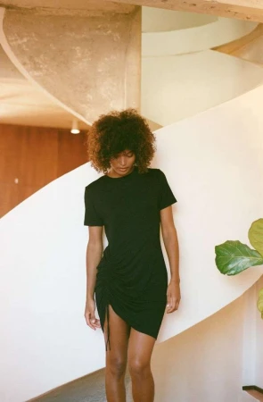 A woman with curly hair poses in a black dress against a spiral staircase, complemented by indoor foliage, creating a fashion-forward look.