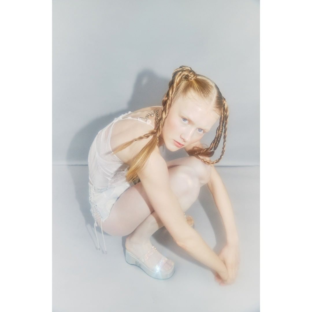 Young woman with blonde braided hair poses in a studio wearing a sheer white dress and platform shoes against a pale grey backdrop.
