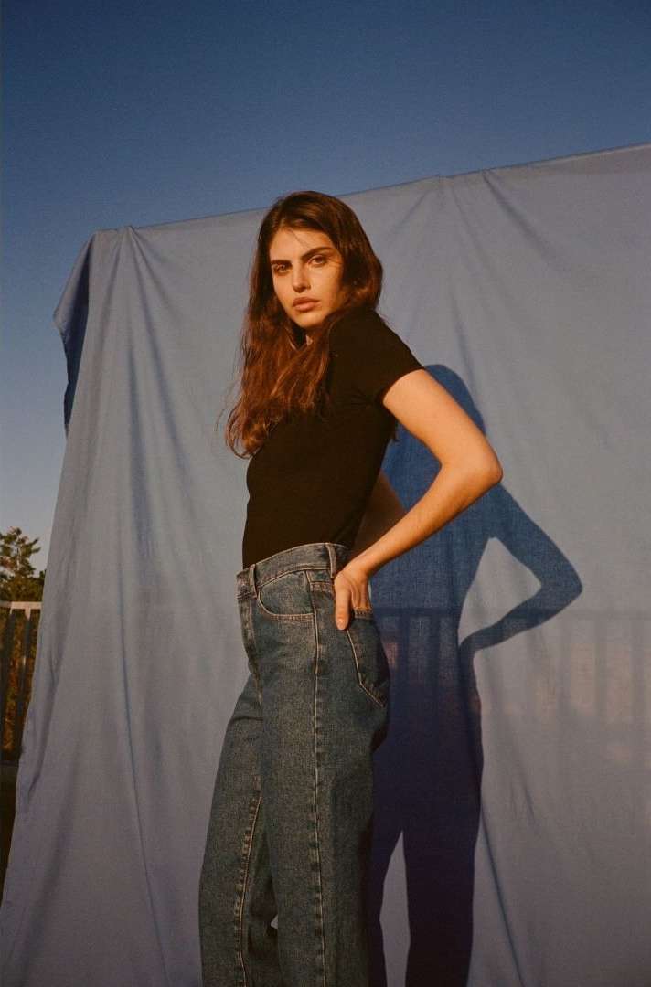 Brunette woman in jeans and black t-shirt poses against a blue backdrop in a medium shot outdoors with natural lighting and soft shadows.