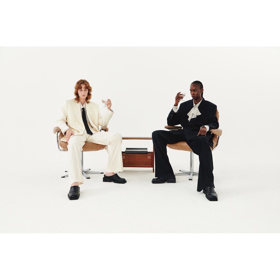 Two people sit in chairs, holding glasses, wearing suits against a white backdrop with a table holding a box between them.