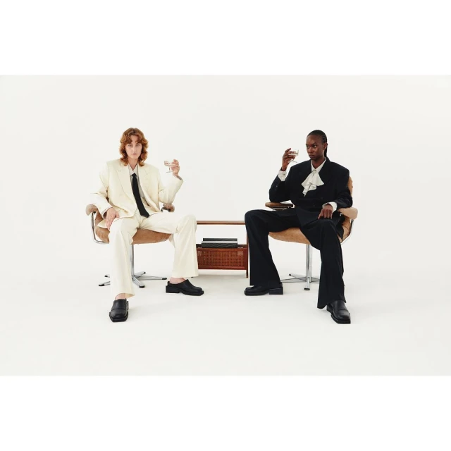 Two people sit in chairs, holding glasses, wearing suits against a white backdrop with a table holding a box between them.
