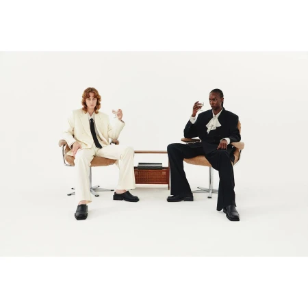 Two people sit in chairs, holding glasses, wearing suits against a white backdrop with a table holding a box between them.
