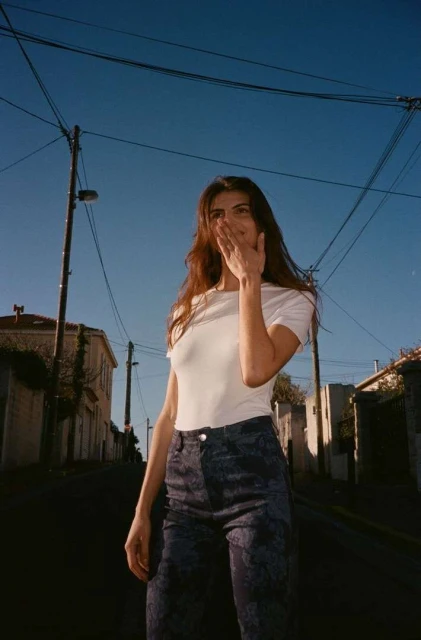 A young woman with long, dark hair smiles and covers her mouth with her hand while standing on a street with buildings and telephone poles.