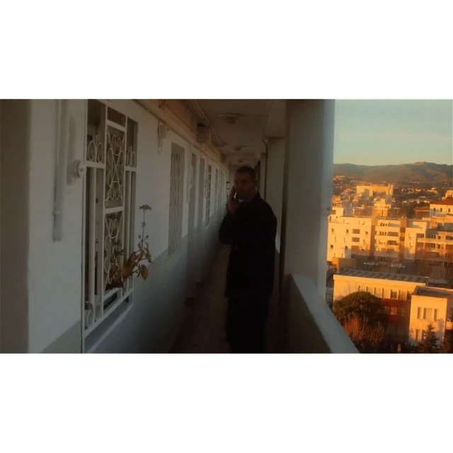 Adult male stands on a white balcony using a mobile phone, cityscape background, and architecture with repeating windows.