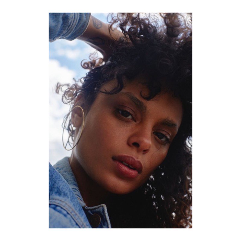 Close-up portrait of a young adult woman with curly dark hair, wearing a denim jacket and large hoop earrings, against a cloudy sky.