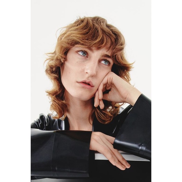 Portrait of a woman with light brown curly hair, wearing a black vinyl top, posing indoors against a white background with a thoughtful expression.