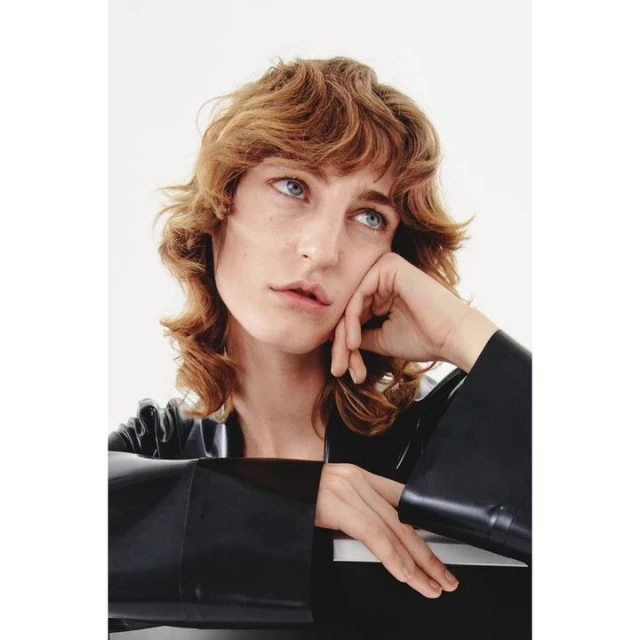 Portrait of a woman with light brown curly hair, wearing a black vinyl top, posing indoors against a white background with a thoughtful expression.