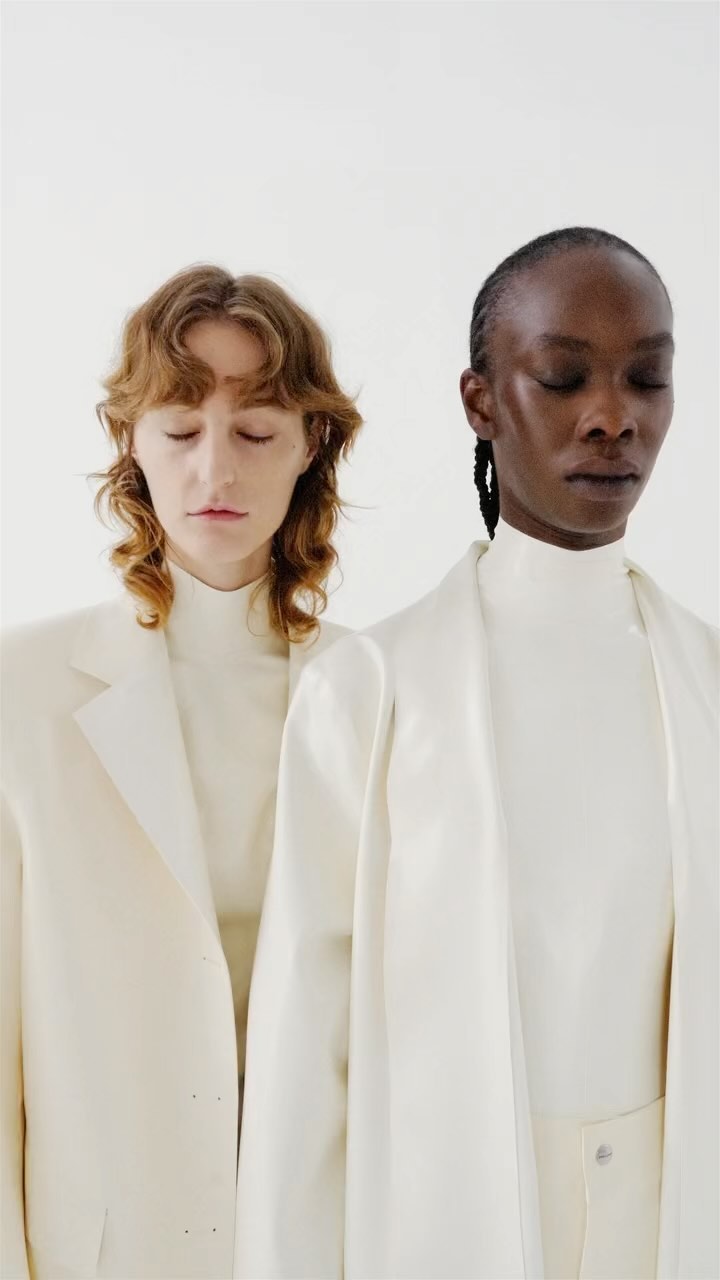 Two fashion models with closed eyes pose in a photo studio wearing elegant, white clothing in a portrait-style, waist-up shot.