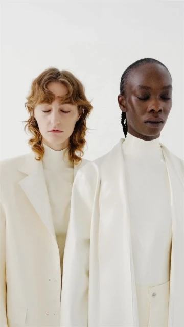 Two fashion models with closed eyes pose in a photo studio wearing elegant, white clothing in a portrait-style, waist-up shot.