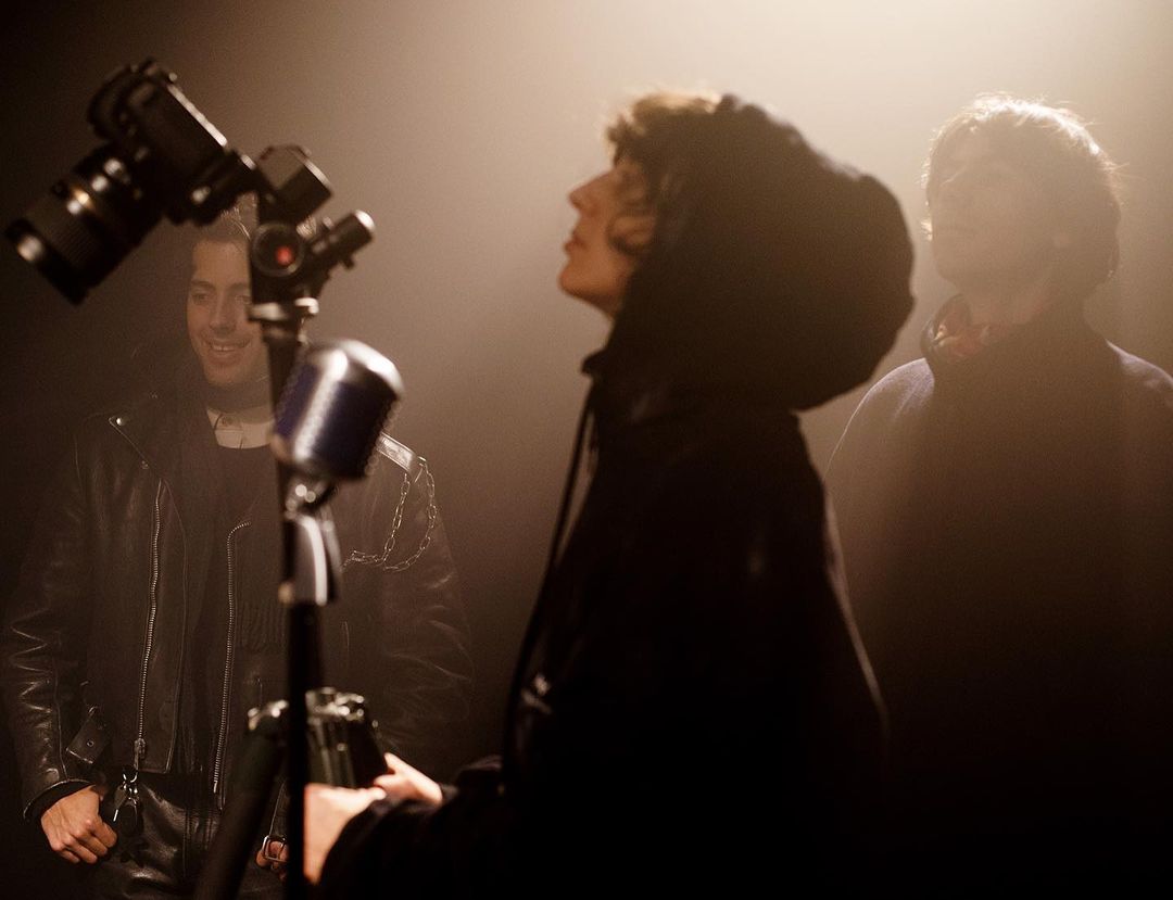 Three men in a music studio setting with vintage microphone, camera, and soft lighting, creating a moody and vintage atmosphere.
