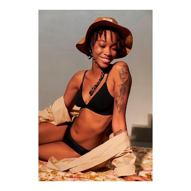 Attractive young woman in a black bikini and hat poses while sitting on a floral-patterned blanket indoors, smiling and relaxed.