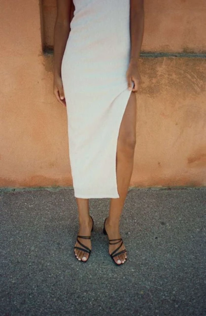 A woman wearing a white dress with a high slit and black strappy sandals stands against a mottled wall on an overcast day.
