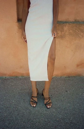 A woman wearing a white dress with a high slit and black strappy sandals stands against a mottled wall on an overcast day.