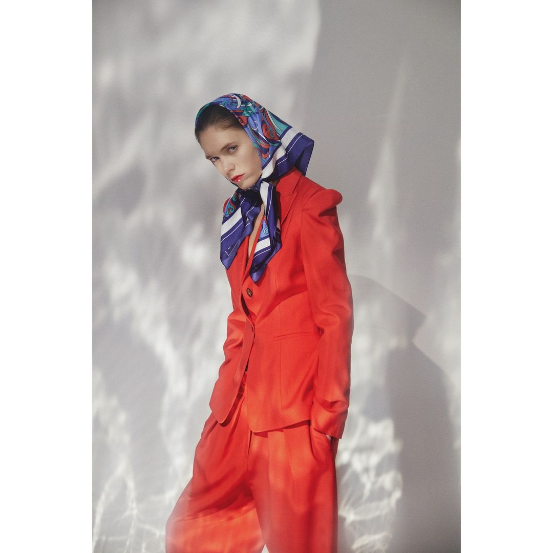 Woman in an orange suit and headscarf poses against a white wall with light reflections. Fashion shot. Studio lit. 