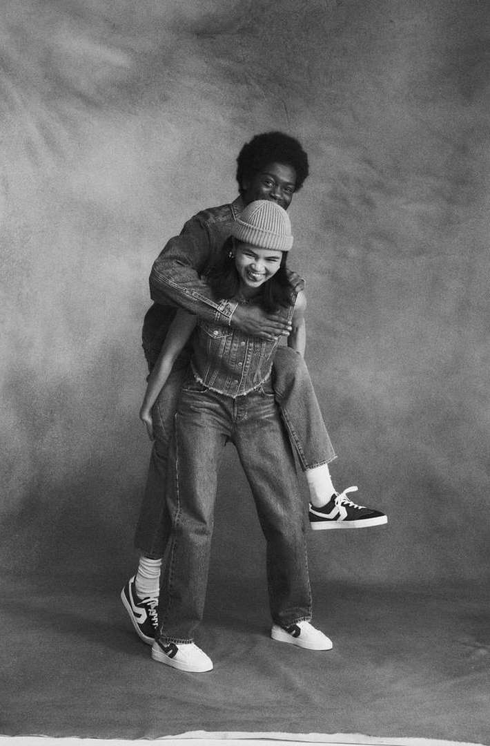 Young woman gets a piggyback ride from a young man in a studio setting. They are both wearing denim and athletic shoes in black and white.
