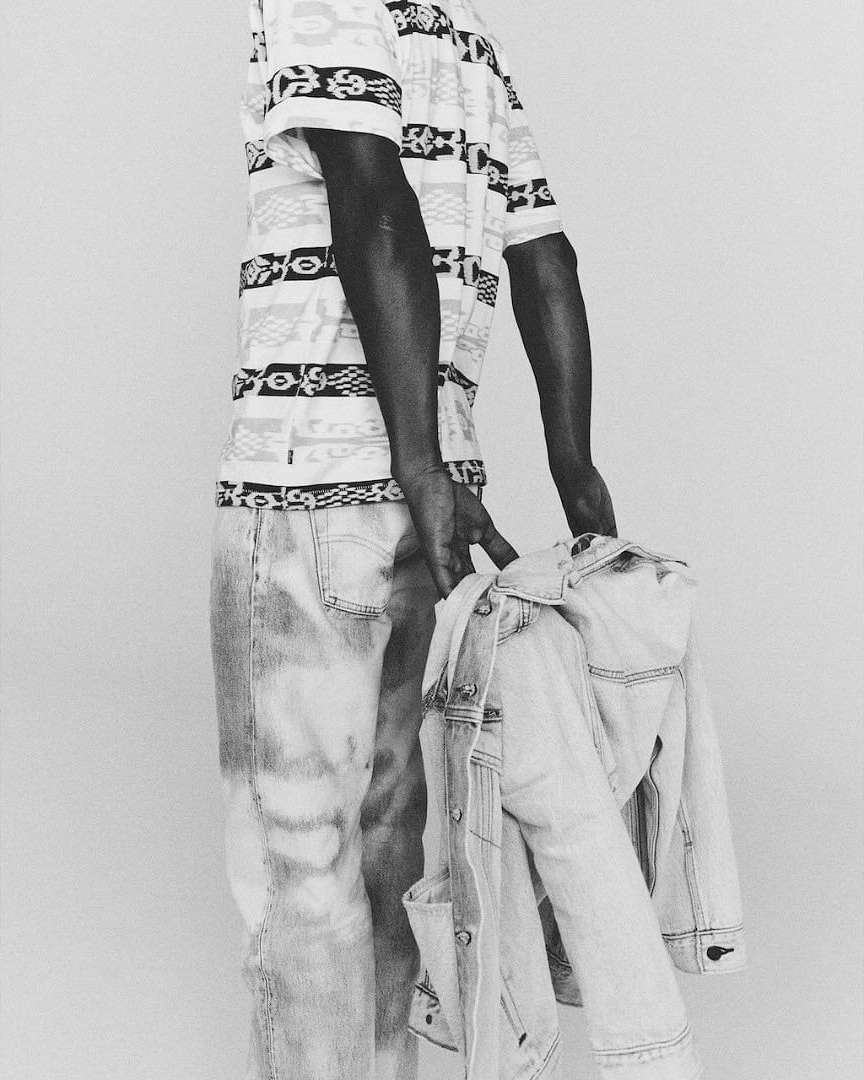 Monochrome portrait of a Black male in patterned shirt and jeans holding a denim jacket in a bright, neutral indoor setting.