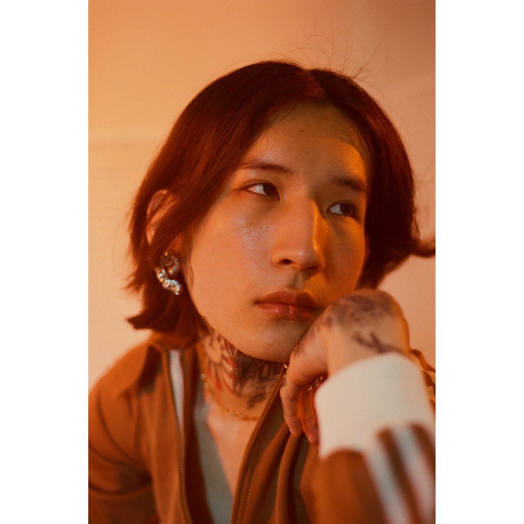 Portrait of a young adult with tattoos, wearing jeweled earrings, and looking off to the side in a warm, golden-toned studio.