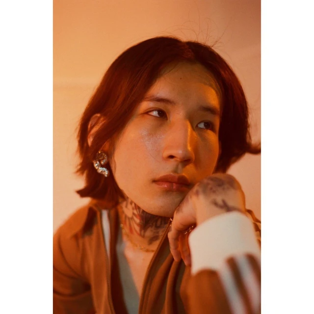 Portrait of a young adult with tattoos, wearing jeweled earrings, and looking off to the side in a warm, golden-toned studio.