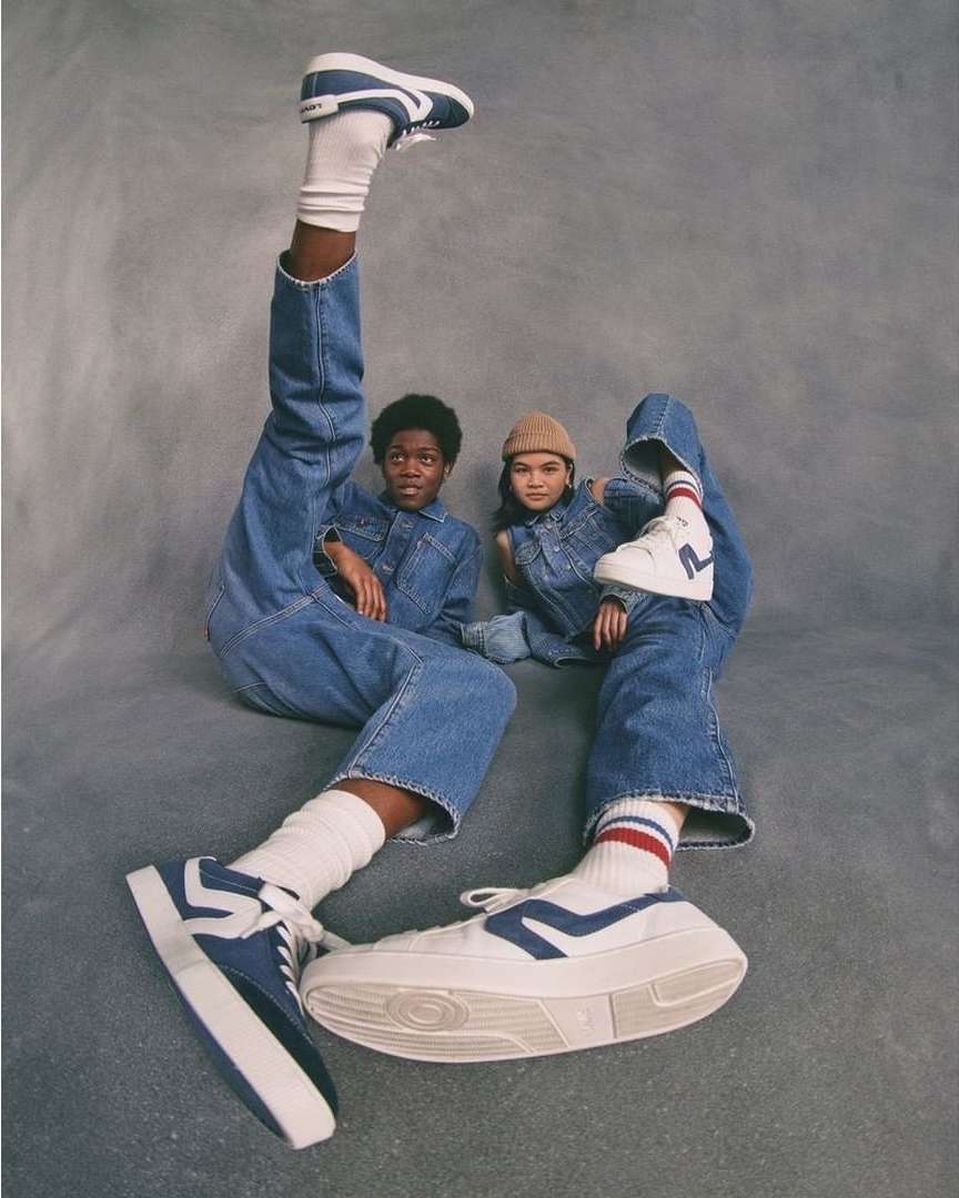 Two children model apparel and white platform sneakers in a studio shot, one with leg raised, against gray backdrop.