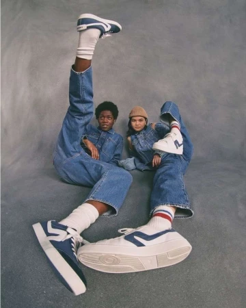 Two children model apparel and white platform sneakers in a studio shot, one with leg raised, against gray backdrop.