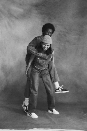 Monochrome portrait of a man giving a piggyback ride to a woman, both in denim outfits and sneakers, set against a mottled backdrop.