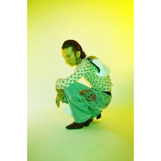 A young adult male crouches in a studio with boxing gloves, wearing a patterned top and jeans, under bright, even yellow lighting.