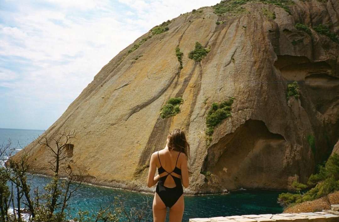 Woman in black swimsuit stands overlooking turquoise sea and rocky coastline with a large rock formation under a cloudy blue sky.