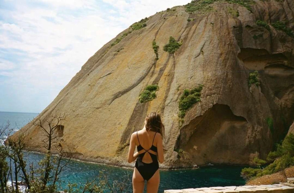 Woman in black swimsuit stands overlooking turquoise sea and rocky coastline with a large rock formation under a cloudy blue sky.