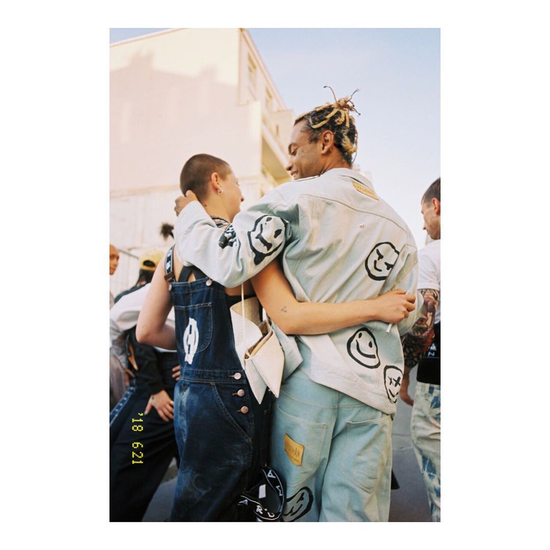 Two people embrace, wearing denim jackets and overalls with smiley face graphics, standing outdoors on a sunny day, fashion-forward.