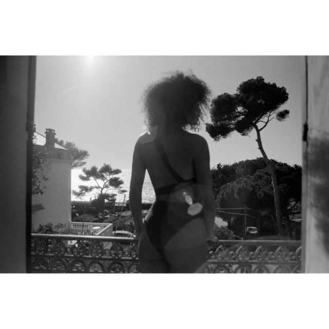 Black and white shot of a woman in swimwear gazing from a balcony toward trees and the ocean on a sunny day, offering a scenic view.