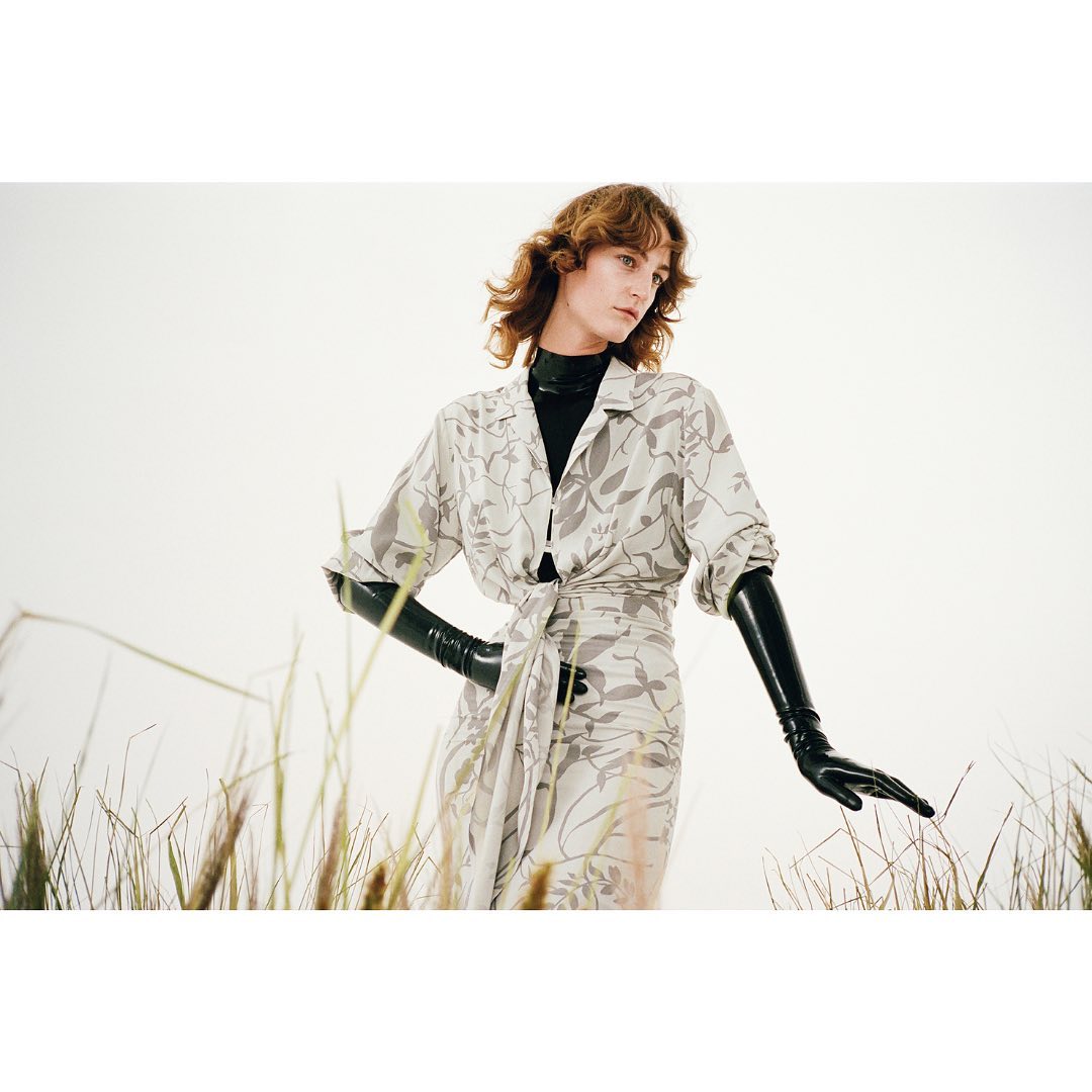Woman in a floral dress posing in a field with dramatic latex gloves, against a white background. Fashion editorial shot.