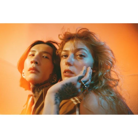 Close-up portrait of two adults with tattoos, one male with long hair, one female with curly hair, posing indoors with vibrant orange lighting.