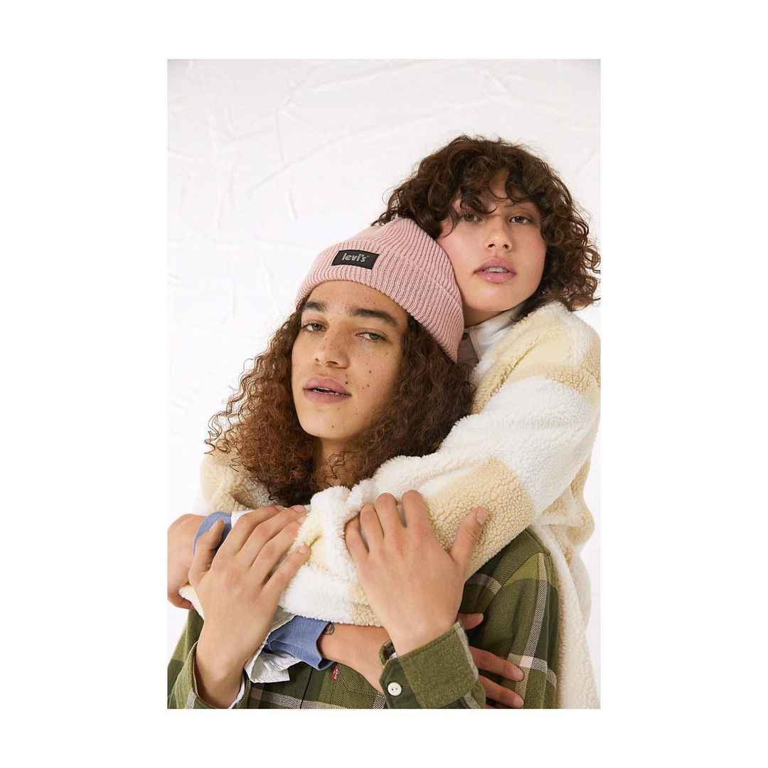 Two young adults embracing against a white background, wearing casual clothing, including a pink beanie and a green plaid shirt.