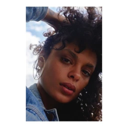 Close-up portrait of a woman with curly dark hair wearing a denim jacket and large gold hoop earrings against a cloudy blue sky.