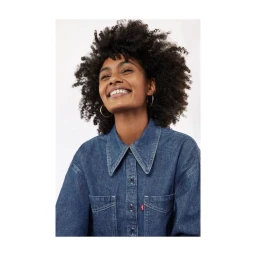 Smiling woman with dark curly hair wearing a blue denim button-up shirt against a plain light background.
