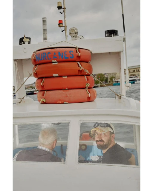 Boat on the water with two adult men on board and stacked orange life preservers on top of cabin against an overcast sky.