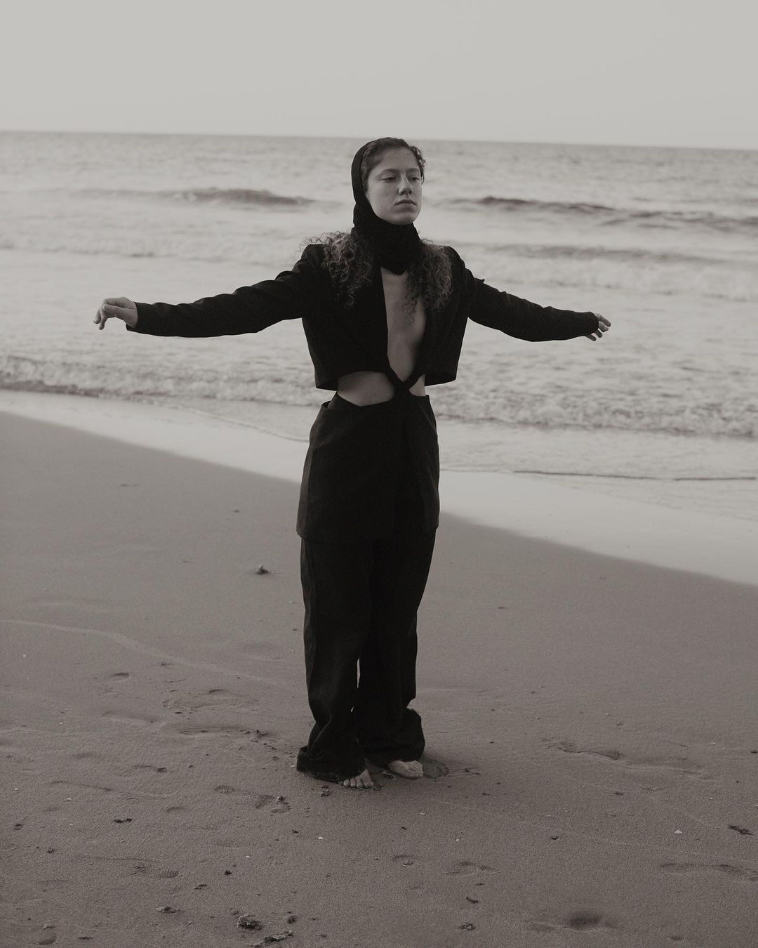 Adult female standing on a beach, arms outstretched, wearing a black cropped jacket and pants with a head covering at sunset.