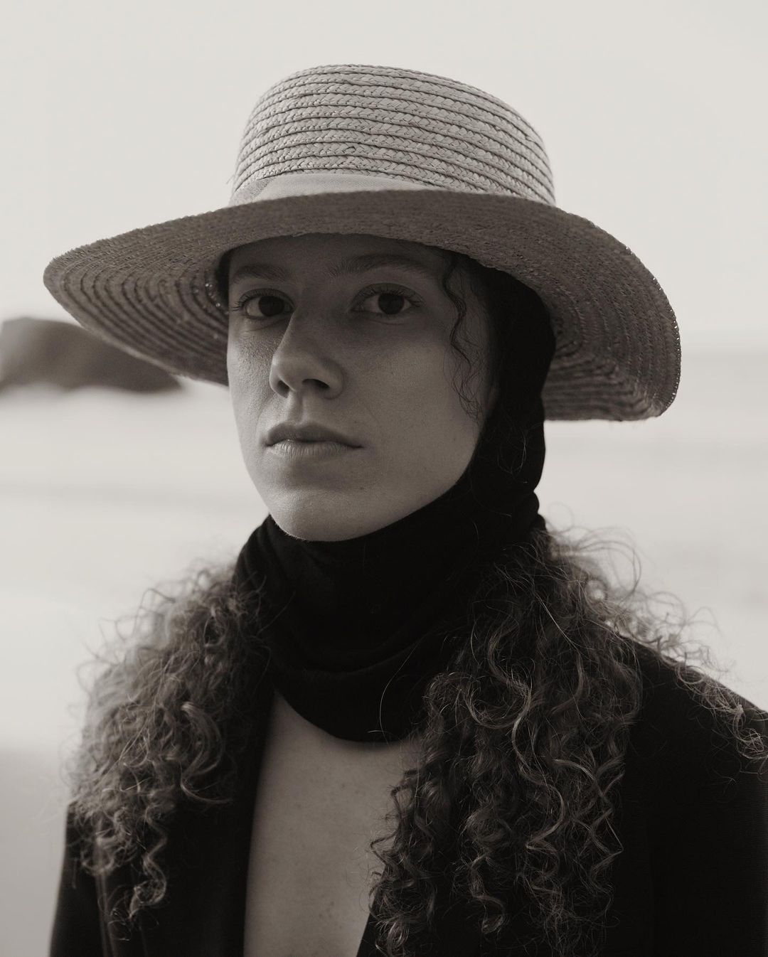 Portrait of a woman wearing a straw hat and dark scarf around her neck against a beach background