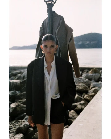 Woman in dark blazer and white shirt standing near a lamp post along a rocky coast in Paris