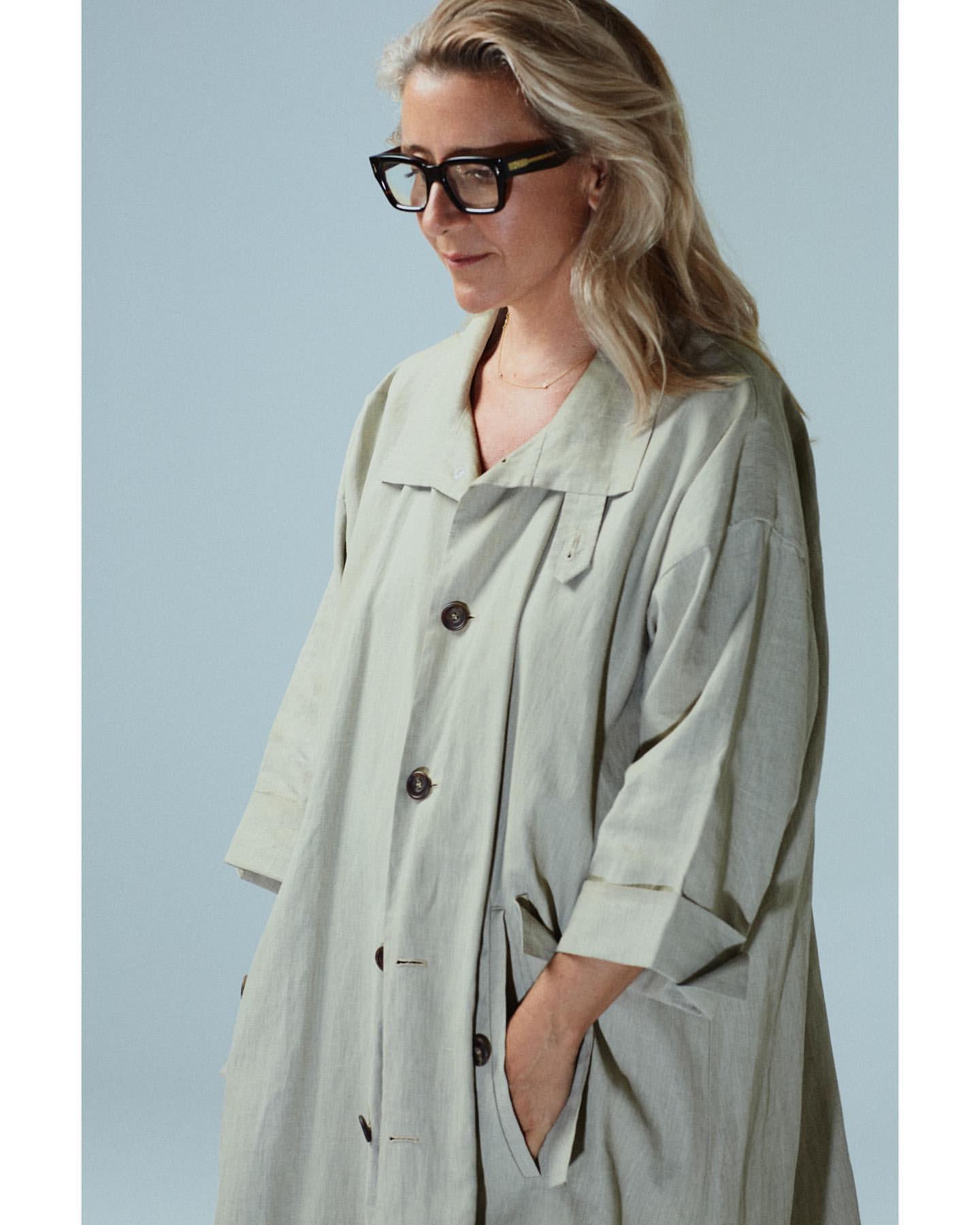 Adult woman in a light-green trench coat poses in a studio with hands in the pocket, wearing black glasses and looking downward.