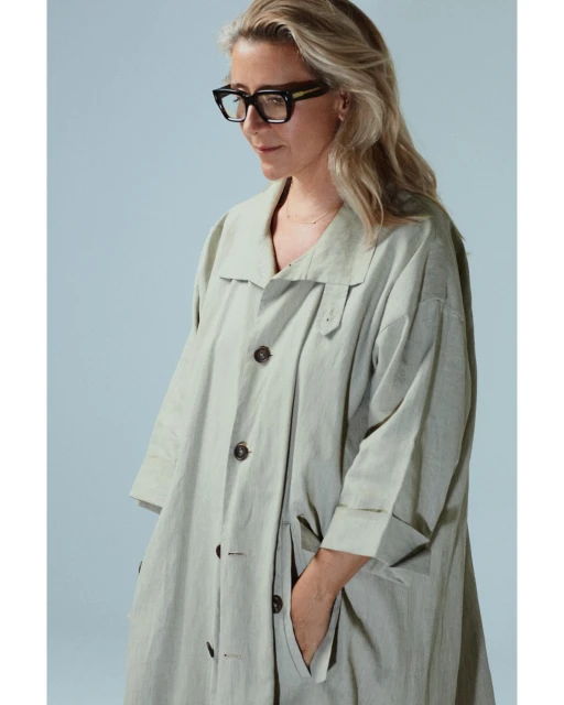Adult woman in a light-green trench coat poses in a studio with hands in the pocket, wearing black glasses and looking downward.