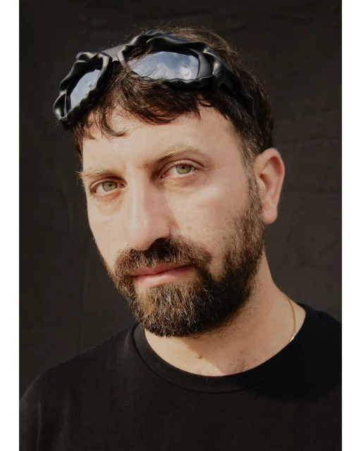 Close-up portrait of an adult male with a beard wearing black goggles against a dark backdrop, showcasing a contemporary style.