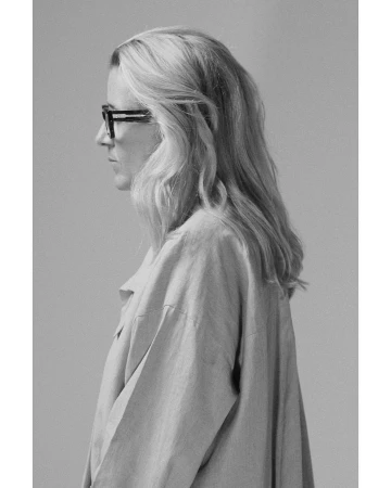 Profile portrait of a blonde woman wearing dark framed glasses and a loose button-down shirt against a neutral grey background.