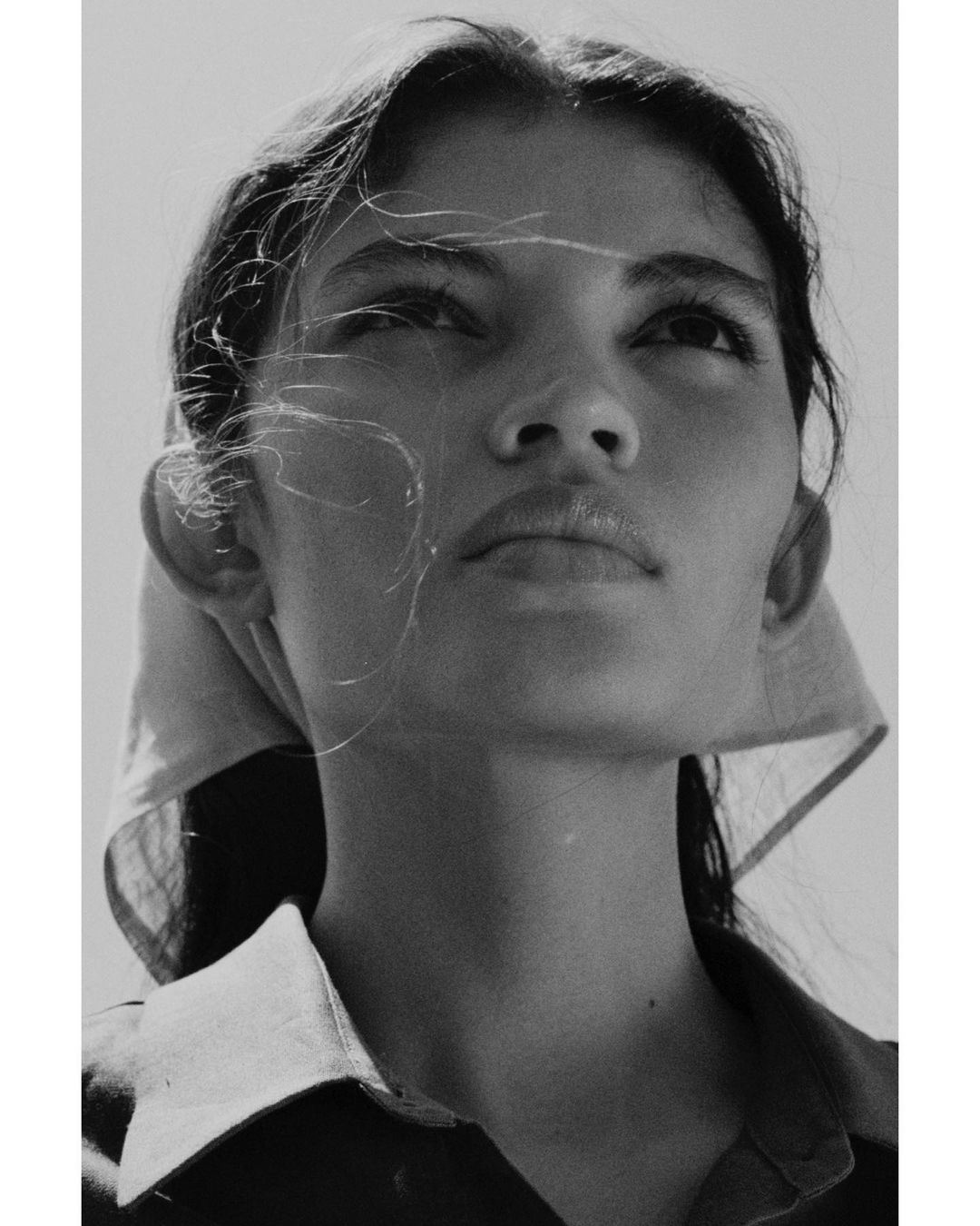 Close-up monochrome portrait of a woman looking upward with wisps of hair blowing across her face, wearing a headscarf.
