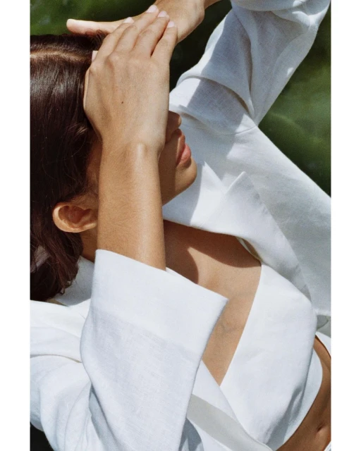 Young woman in white linen outfit poses in sun, shielding face with her hand against a green background. Fashion and summer concept.