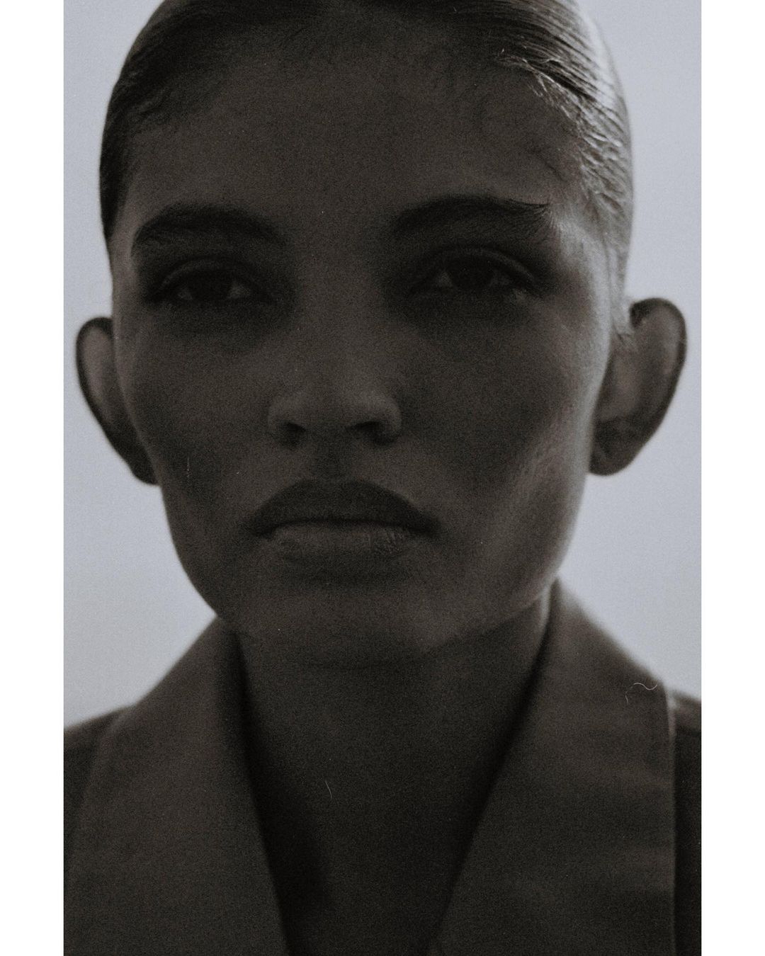 Monochromatic portrait of an adult woman with slicked-back hair wearing a collared shirt against a neutral, soft gradient background.