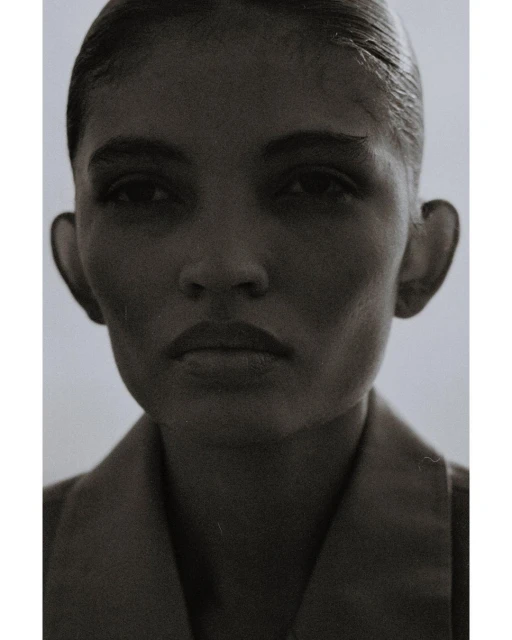 Monochromatic portrait of an adult woman with slicked-back hair wearing a collared shirt against a neutral, soft gradient background.