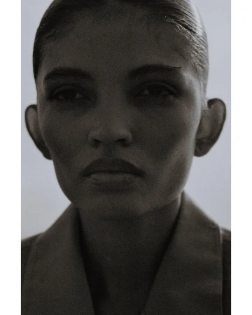 Monochromatic portrait of an adult woman with slicked-back hair wearing a collared shirt against a neutral, soft gradient background.