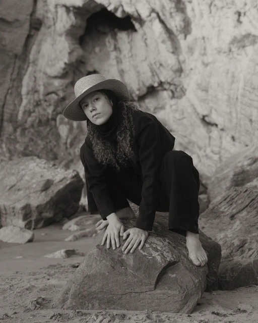 Monochrome portrait of a young woman with curly hair wearing a straw hat and dark clothing, crouching on a rock with a rock cave behind.